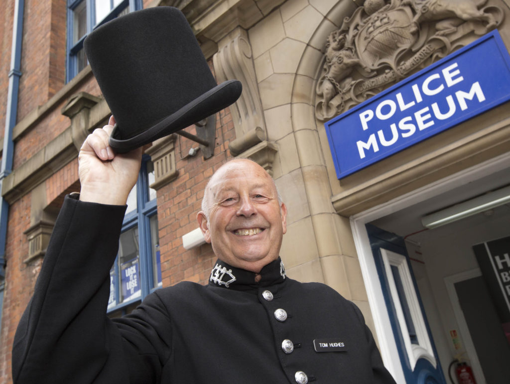 A smiling man dressed as a 19th Century policeman raises his top hat outside the front entrance of the Police Museum