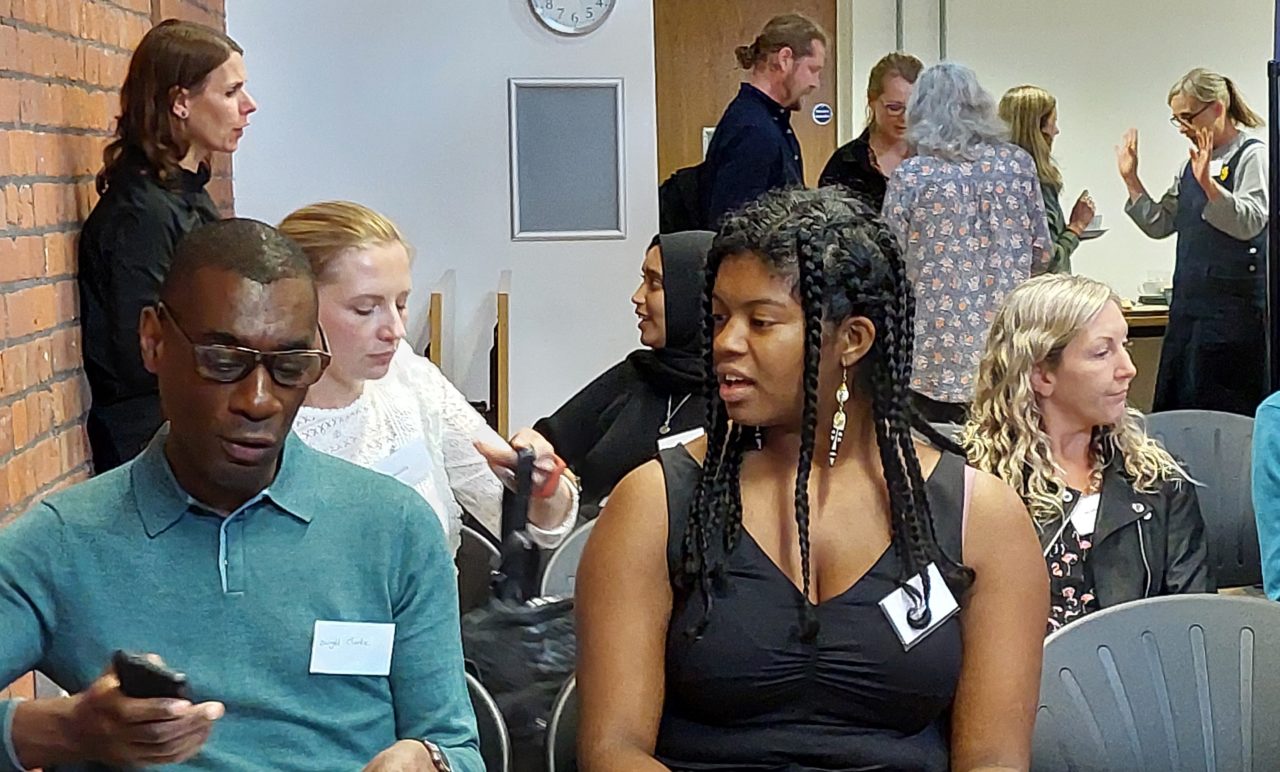 A man holding a mobile phone and a woman are talking to each other, sitting down at a conference with other people standing in the background.