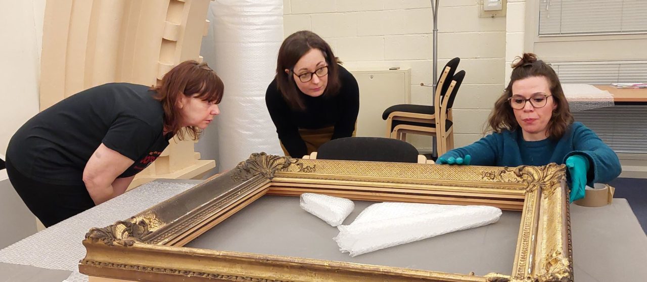 Three women at a Museum Basics training looking at the back of a framed picture on a table to prepare for packing it into tissue paper.