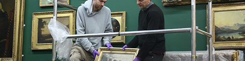 Three men working in a museum gallery with paintings on the wall behind them. A third man wearing gloves passes a small painting to the others.