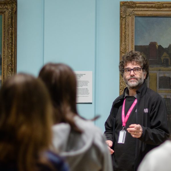A museum assistant wearing a lanyard stands in a gallery in front of a painting and talks to an audience of young people.