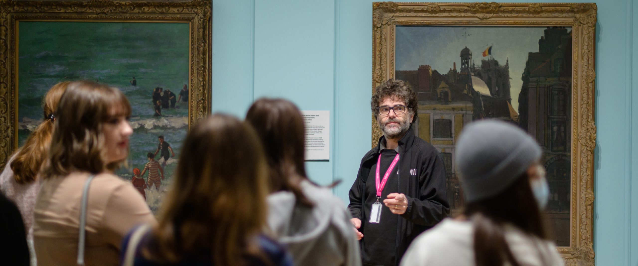 A museum assistant wearing a lanyard stands in a gallery in front of a painting and talks to an audience of young people.
