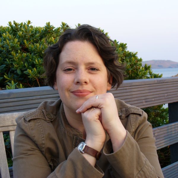 A woman with dark hair and a khaki jacket sitting on a bench with a hedge and the sea behind her.