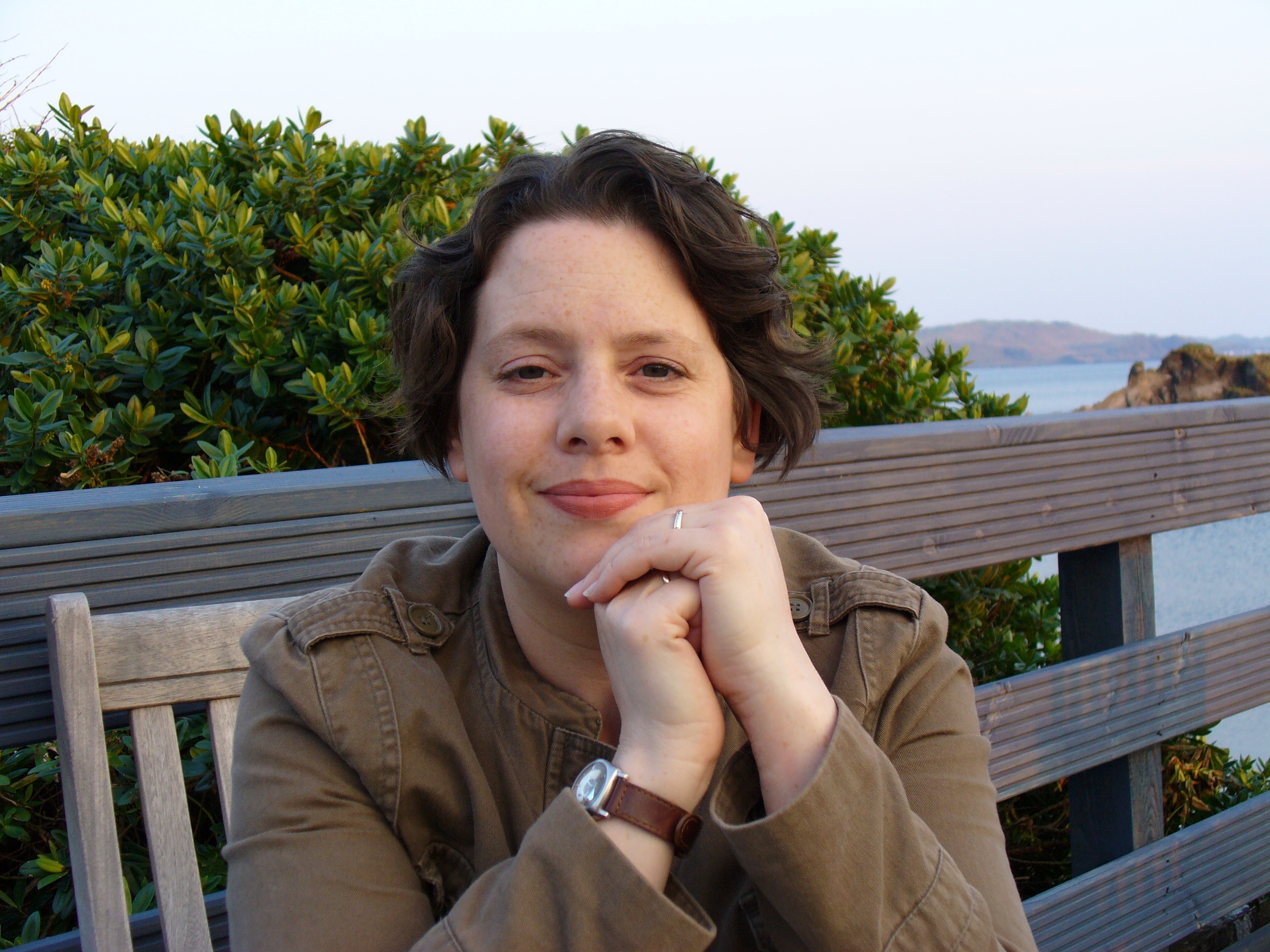 A woman with dark hair and a khaki jacket sitting on a bench with a hedge and the sea behind her.