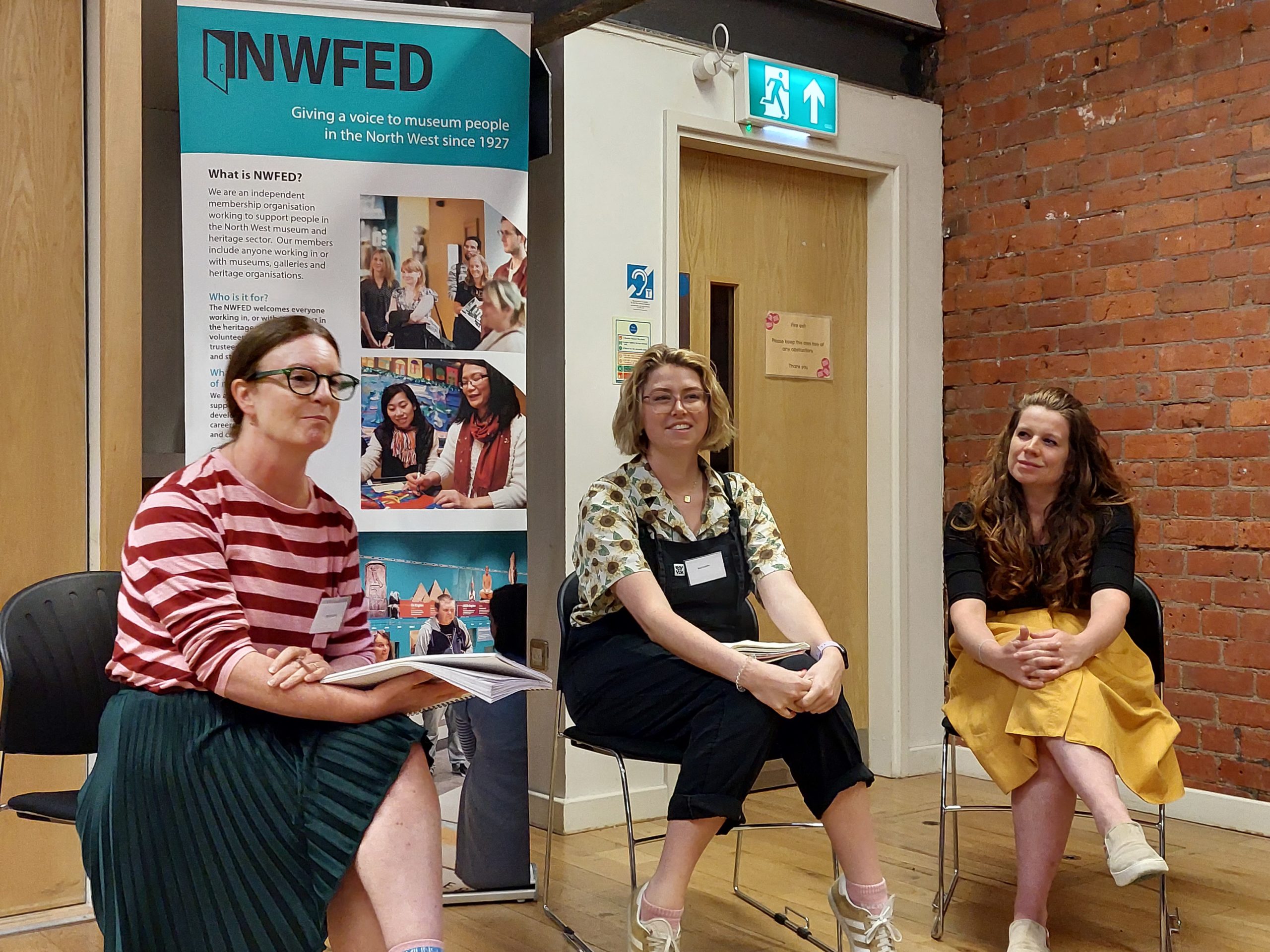 Three female staff members from Imperial War Museum North West and Manchester Jewish Museum sitting in a row in front of a NWFed banner.