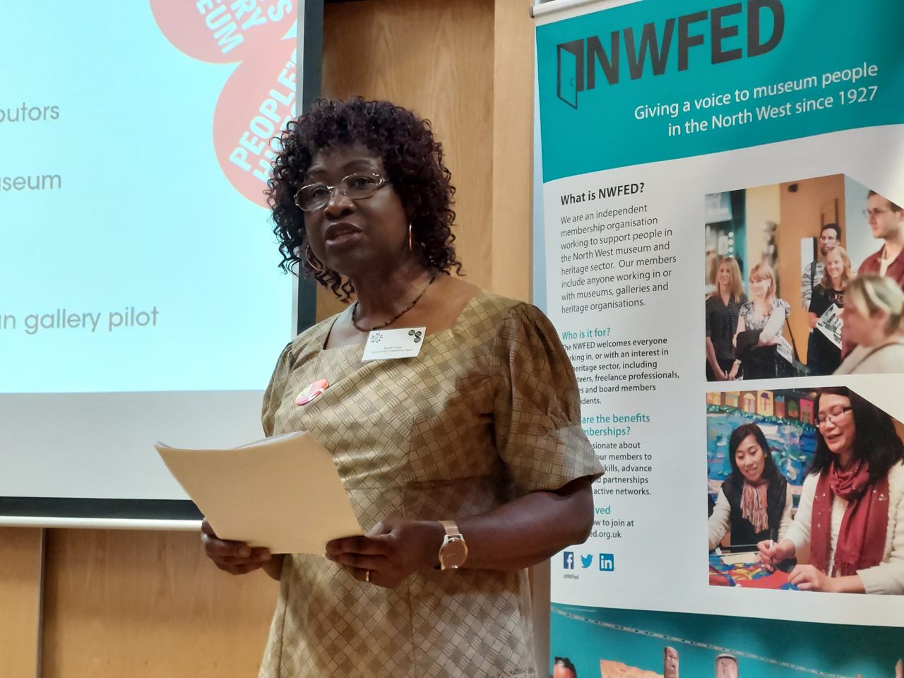 Agnes Fough, a member of the People's History Museum Community Panel, stands in front of a screen holding a piece of paper. Behind her is the NWfed logo