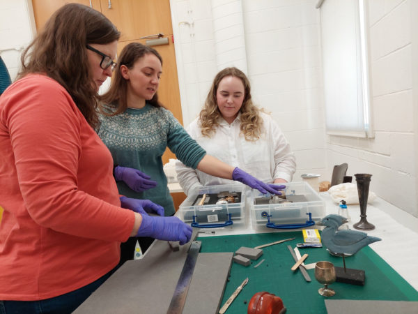 Three women wearing latex gloves using Plastazote and plastic boxes to safely pack small, metal museum objects on a table.
