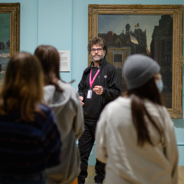 A museum assistant wearing a lanyard stands in front of a painting talking to an audience of young people standing in the gallery