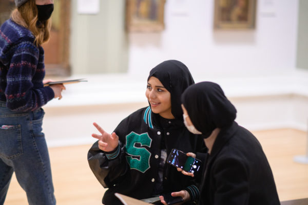 Two young Muslim women sitting and talking in a museum gallery with paintings on the wall.