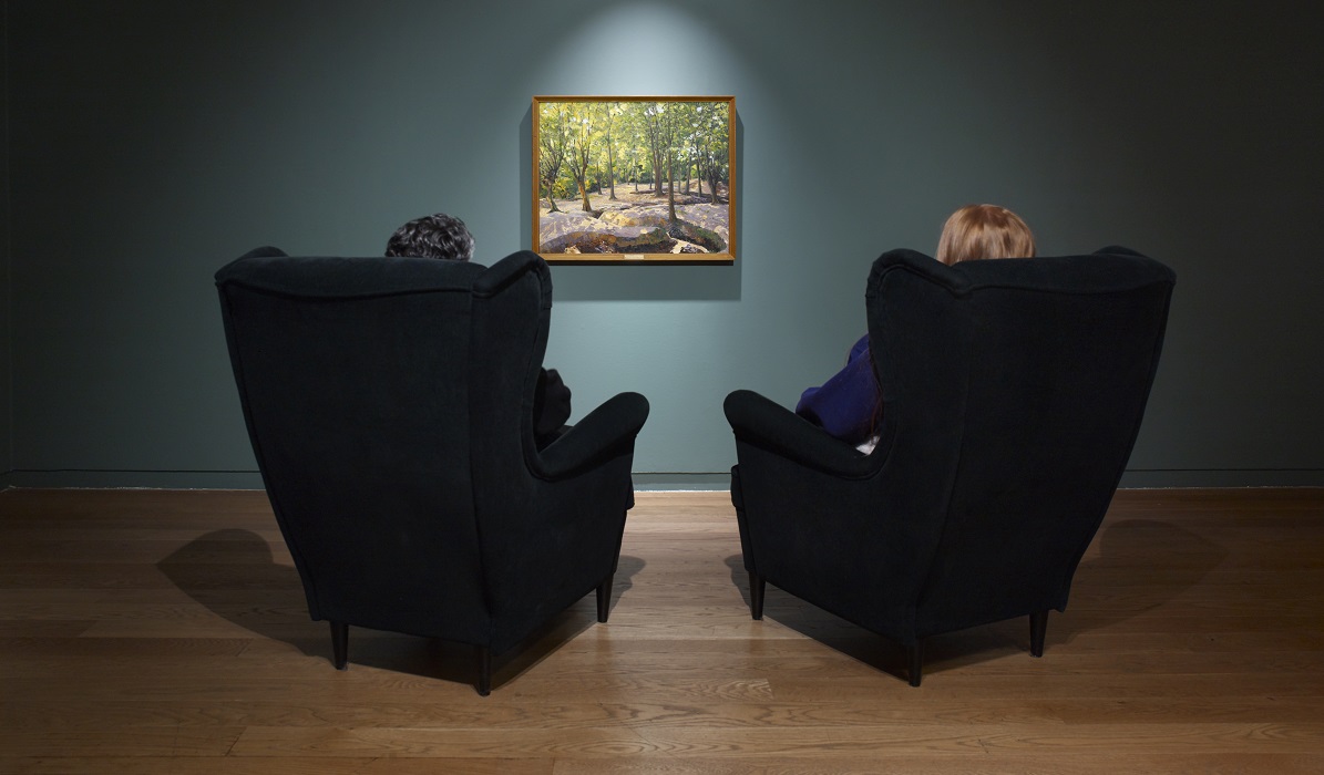 Two people sat on armchairs looking at a framed painting on a blue gallery wall